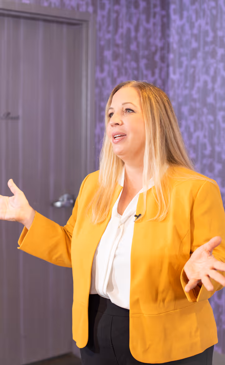 Woman with long blonde hair wearing a yellow blazer and white blouse speaking with arms outstretched.