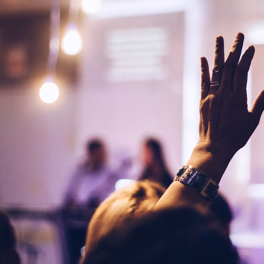Close-up of a person raising their hand in a dimly lit room with two blurred figures in the background.