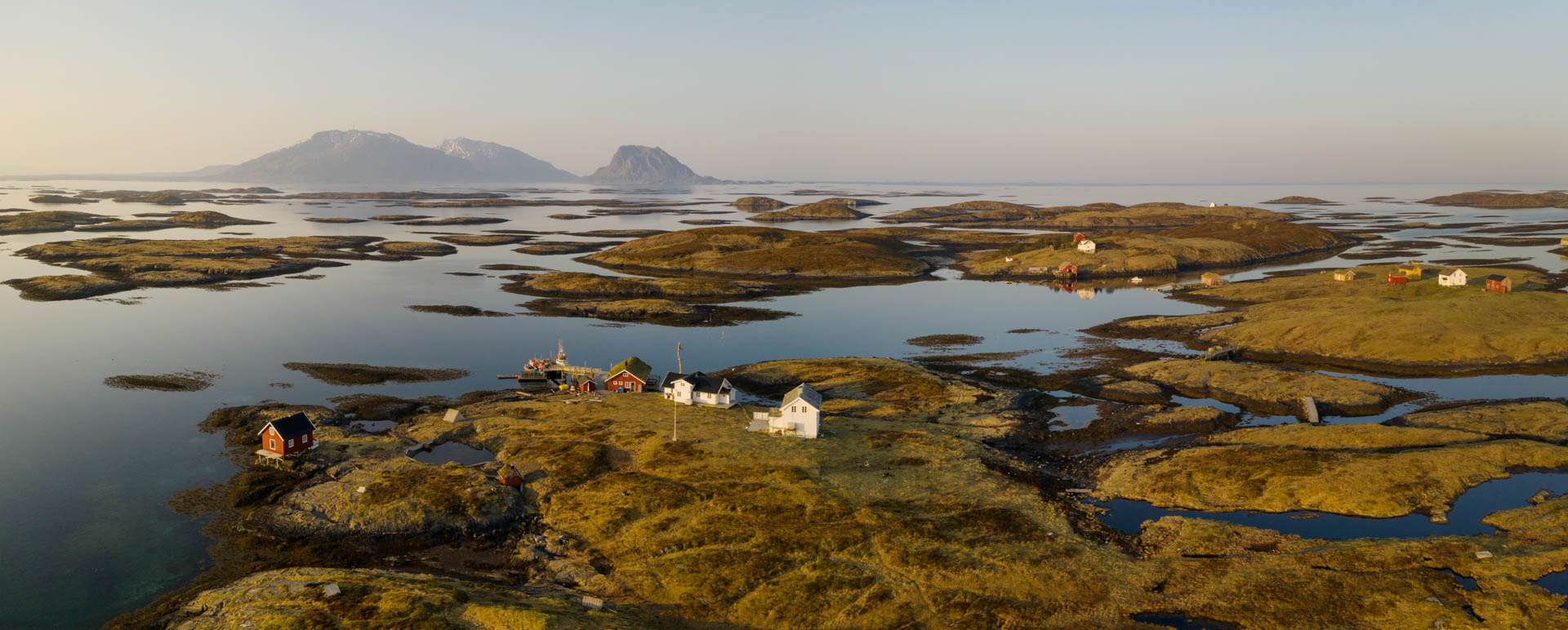 Flere mindre gårder er spredt utover småøyene i Vegaøyan. Havet er blikkstille og i bakgrunnen ses øya Vega.