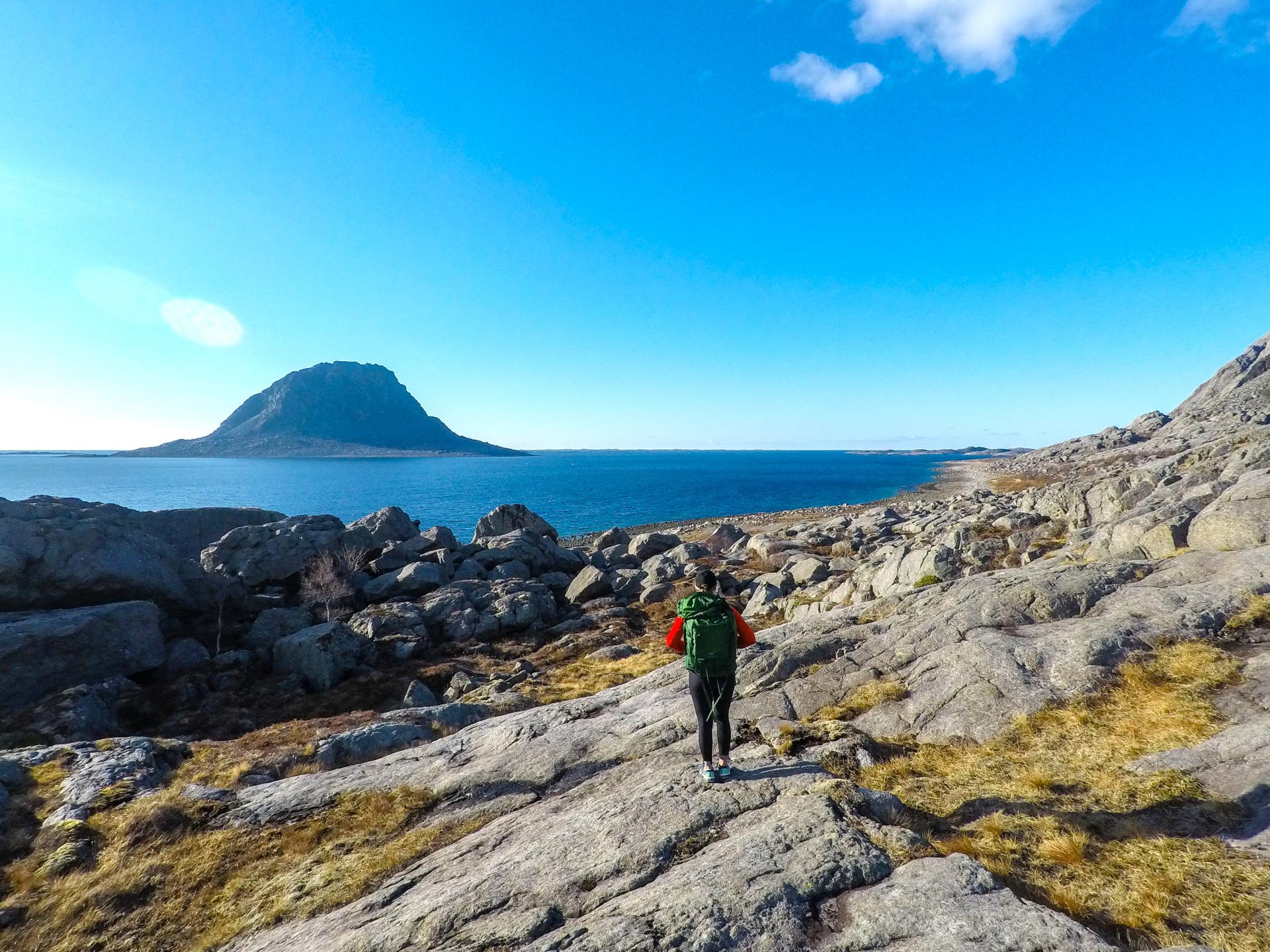En turgåer som går langs blankskurte berg langs kysten av øya Vega. I bakgrunnen ses en rullesteinsstrand og fjelløya Søla.