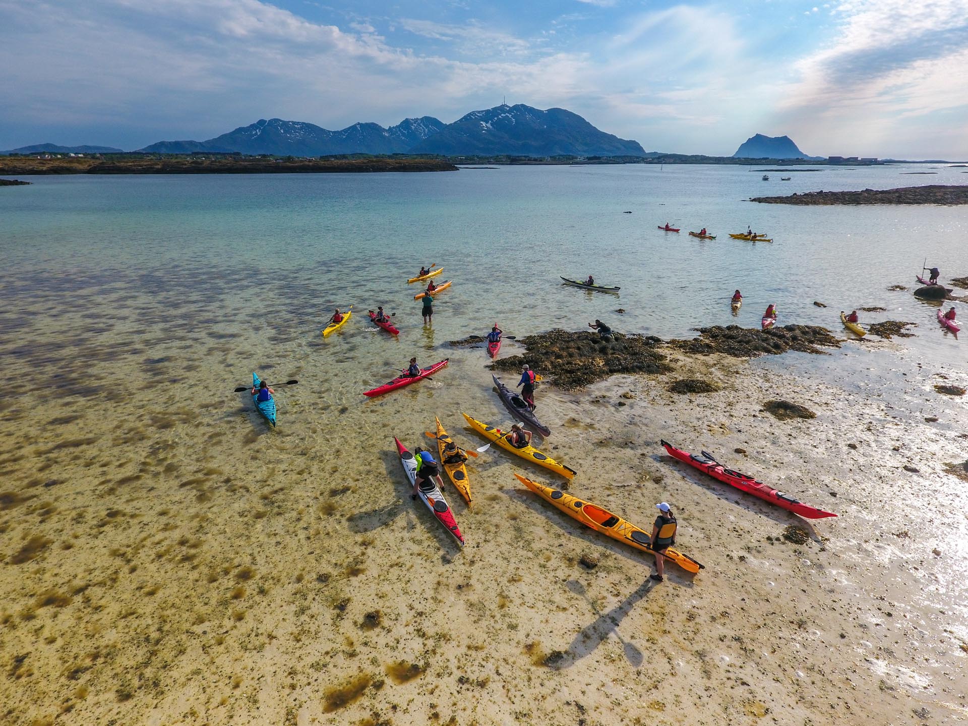 Flere kajakker er på vei ut fra en langgrunn sandstrand. Solen skinner og i bakgrunnen ligger øya Vega.