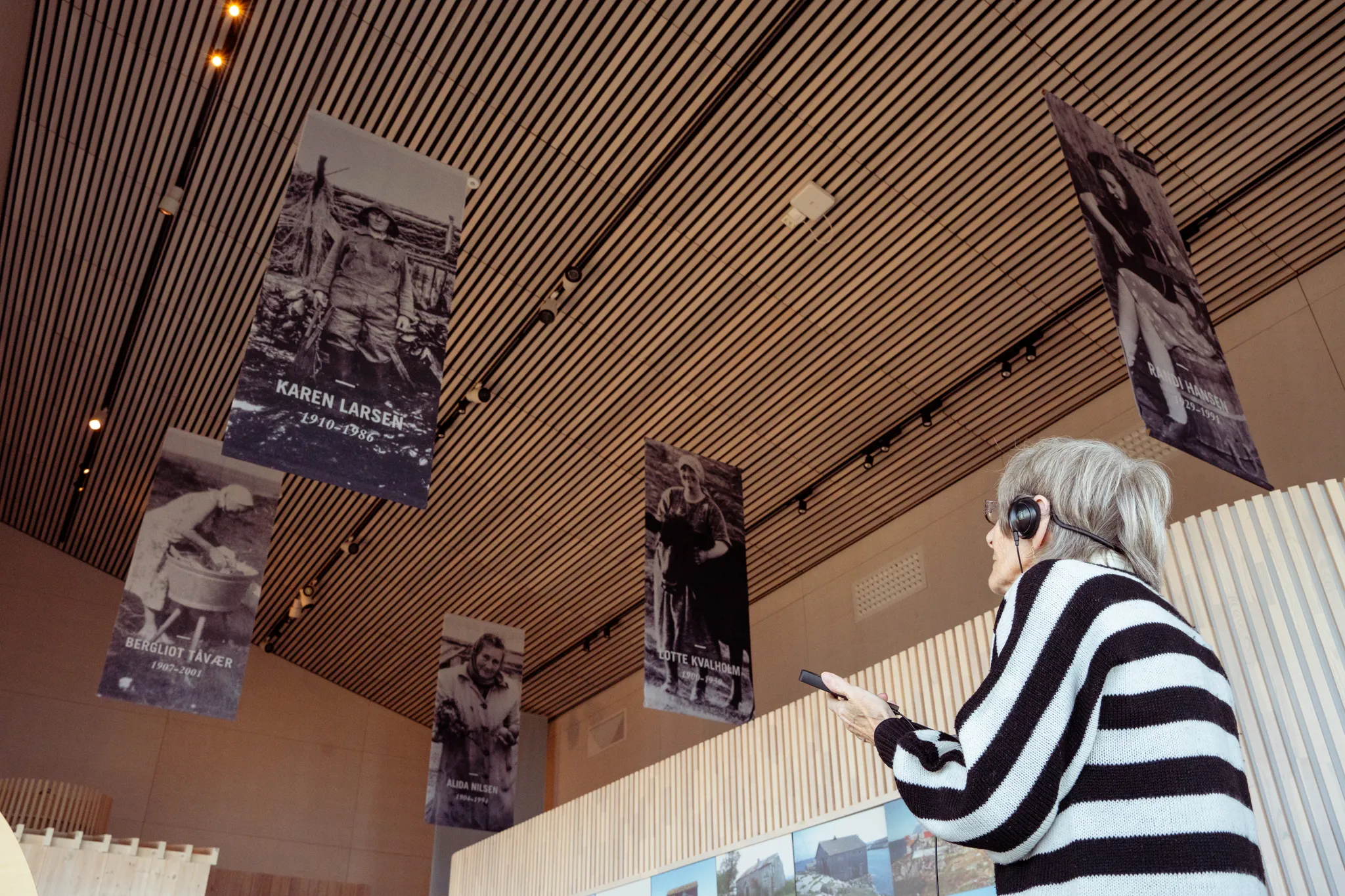 En gjest står i utstilingen Ville vakre Vega og lytter til lydhistorier på en audioguide. I taket over henne henger det store svarthvitt bilder av ærfuglvoktere.