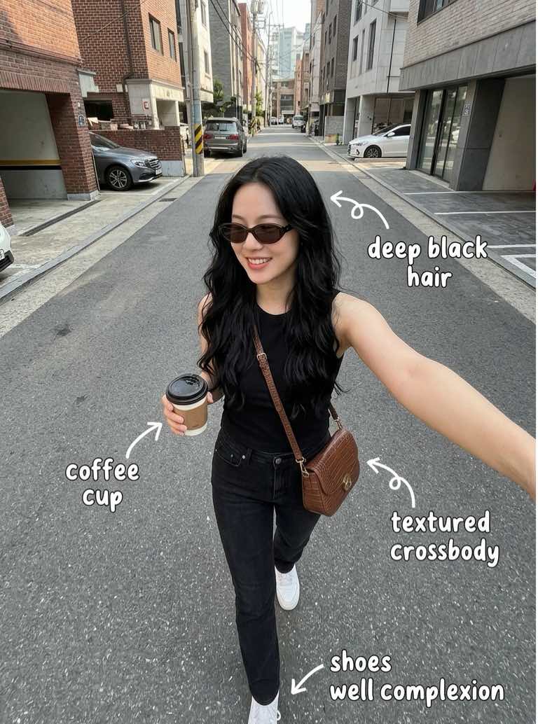 Woman with long black hair walking down a city street holding a coffee cup while wearing sunglasses and a brown crossbody bag.