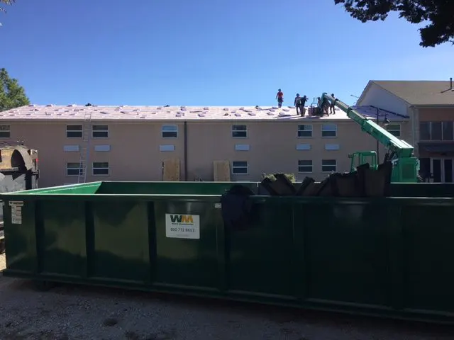 Roofing crew installing new commercial roof on multi-story building with green waste container on site.