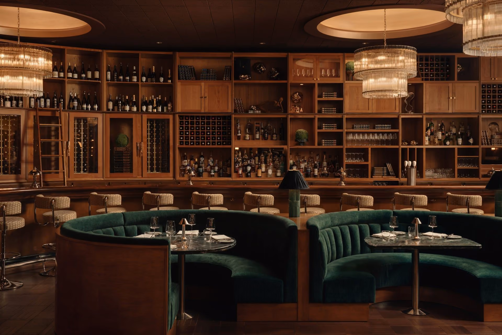Elegant bar with dark green velvet curved booths, marble tables, wood shelves, and warm lighting chandeliers.