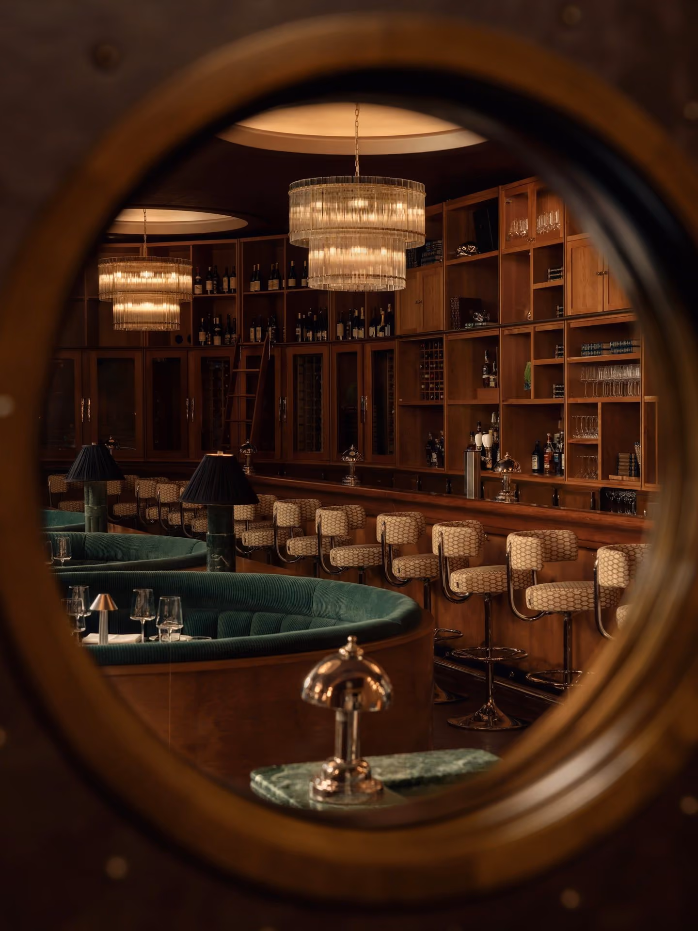 View of a vintage bar with green seats, patterned bar stools, chandeliers, and wooden shelves with bottles.