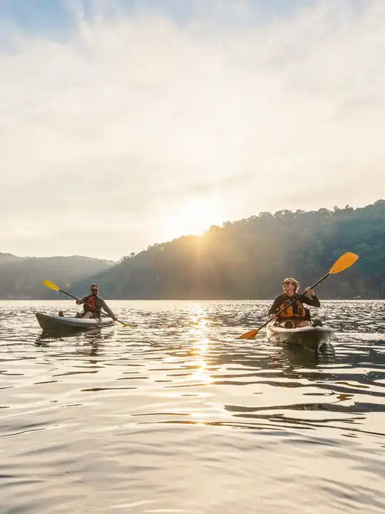 Two people kayaking on a calm lake with hills in the background and the sun shining through partly cloudy sky.