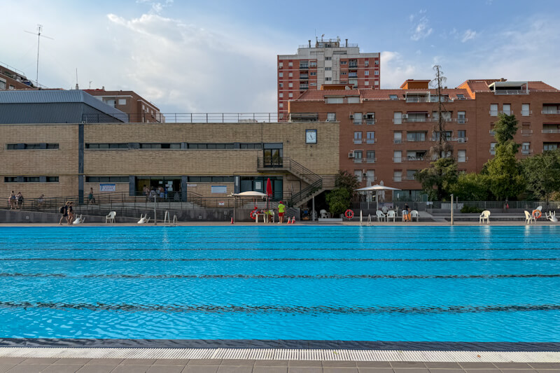 Outdoor swimming pool at Canal de Isabel II Sports Centre in Madrid with lap lanes, pool deck, and residential buildings in the background.