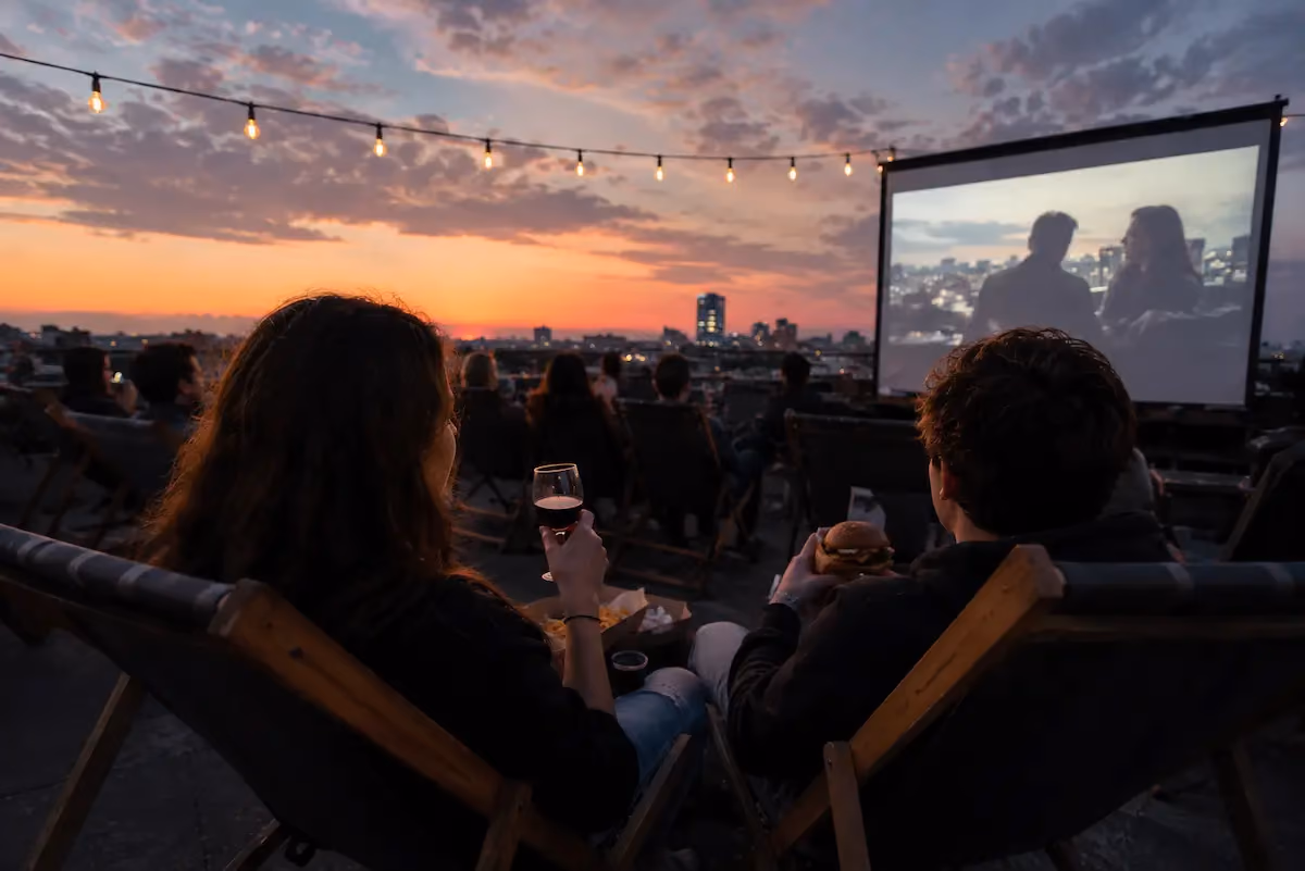 Un grupo de amigos viendo una película en un cine al aire libre en una azotea durante el atardecer, con el horizonte de la ciudad de fondo.