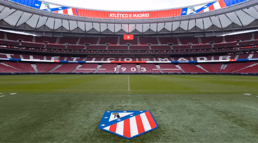 Panoramic view of Atlético de Madrid's stadium from pitch level, featuring the club crest on the grass and the iconic "1903" seating display in the stands.