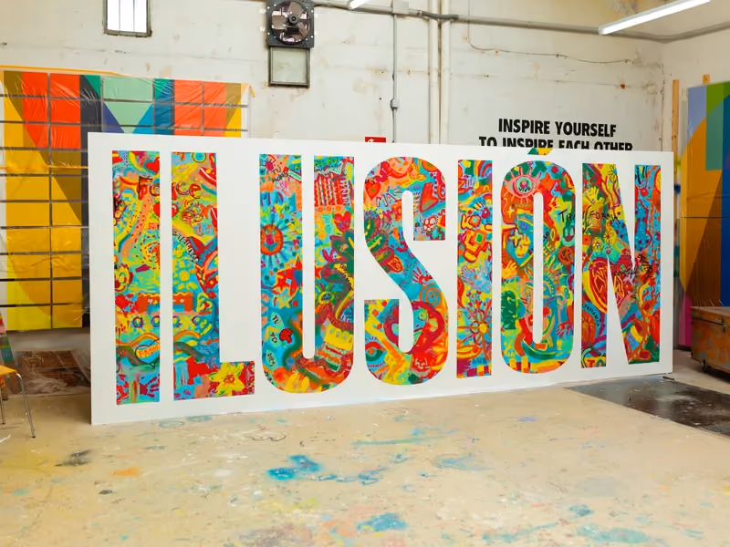 Large freestanding letters spelling ILUSIÓN, covered in colourful collaborative artwork, in a studio.