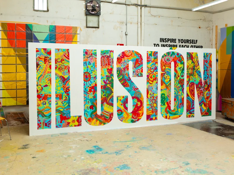 Large freestanding letters spelling ILUSIÓN, covered in colourful collaborative artwork, in a studio.