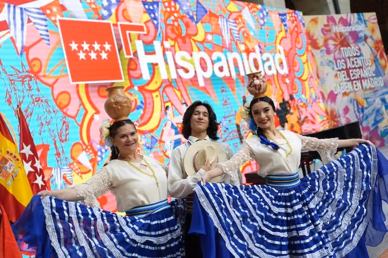 Dancers in traditional dress in front of a colorful screen reading "Hispanidad" for Madrid's October 12th celebration.