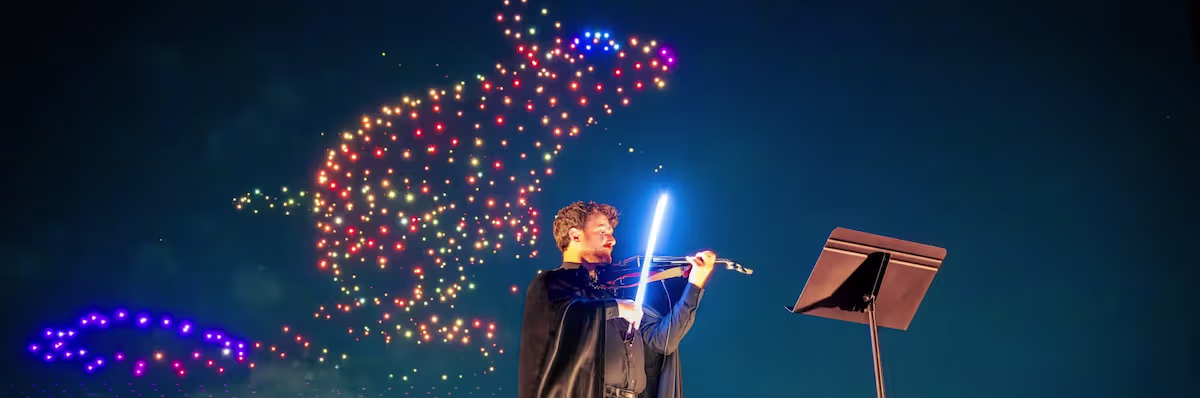 Violinist playing under glowing lights as drones form a Harry Potter figure in the night sky.