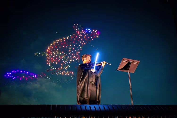 Violinist playing under glowing lights as drones form a Harry Potter figure in the night sky.