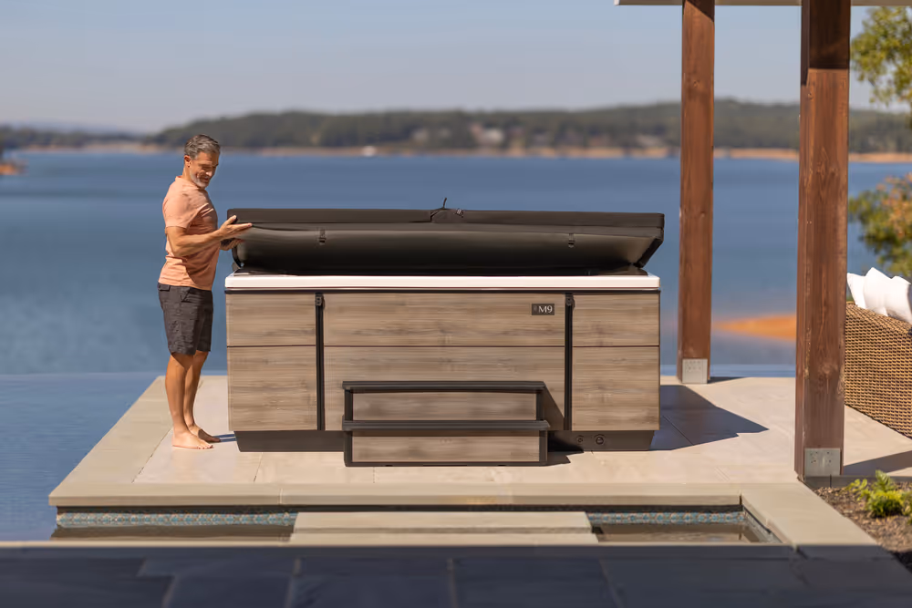A man removing the cover of an M9 spa with scenic outdoor backdrop.