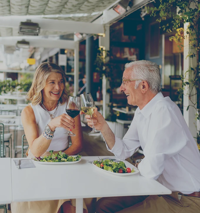 Smiling retired couple toasting with wine glasses while seated at a restaurant table with salad plates.