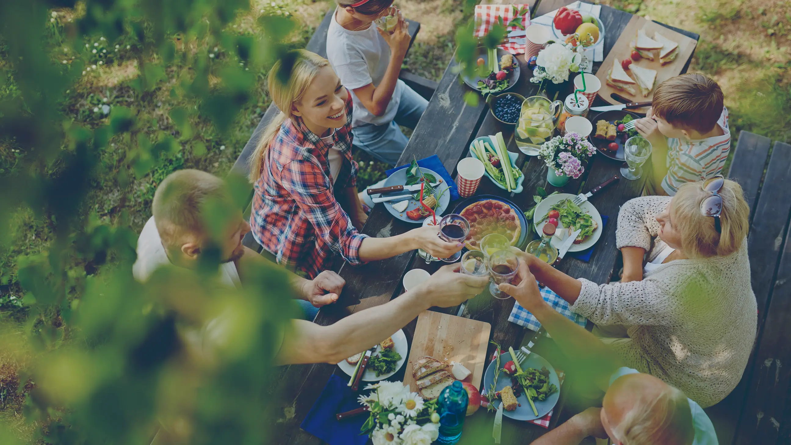 Family gathered around a picnic table outdoors, toasting with glasses over a meal including pizza, vegetables, and sandwiches.