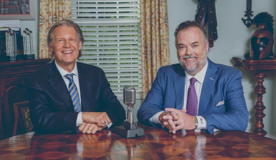 Phillip Allen and Tony Dale from Common Sense Retirement Planning in suits smiling and seated at a polished wooden table with a vintage microphone between them in a room with bookshelves and curtains.