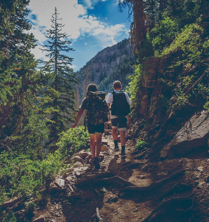 Two hikers with backpacks walking on a rocky forest trail surrounded by trees and mountains under a partly cloudy sky.