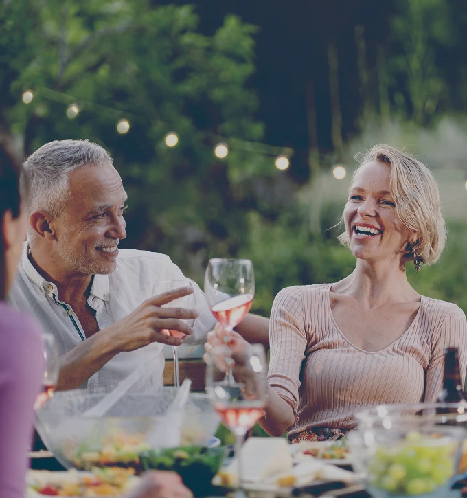 Smiling man and woman clinking wine glasses during an outdoor dinner party with food and string lights in the background.