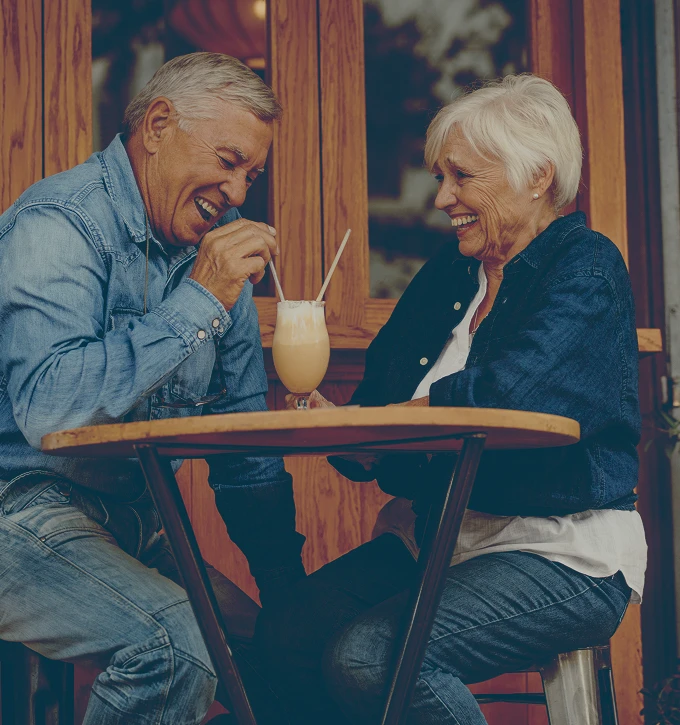 Retired couple laughing and sharing a drink with two straws at an outdoor table.