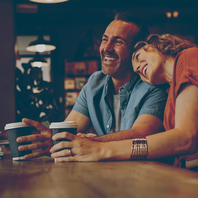 Smiling man and woman sitting at a table in a café holding takeaway coffee cups.