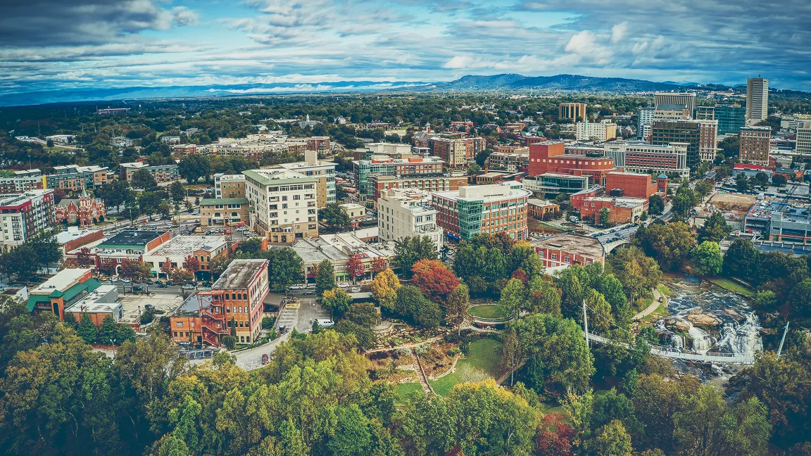 Aerial view of Greenville, South Carolina cityscape featuring mixed buildings, lush green trees, and a river with a small waterfall and pedestrian bridge under a partly cloudy sky.