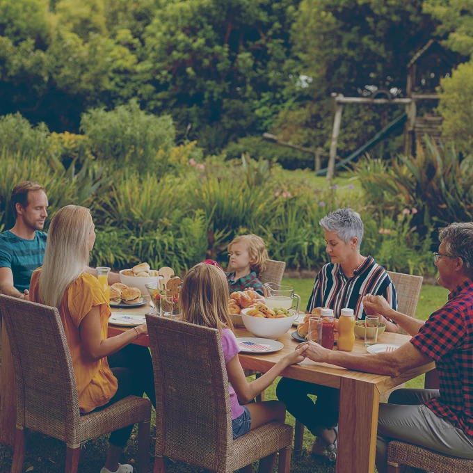 Family of six holding hands and praying around a wooden outdoor table set with food in a garden.