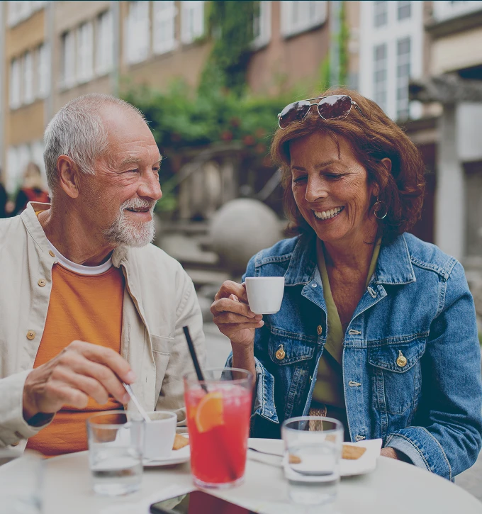 Retired couple enjoying coffee and a pink beverage at an outdoor café table, smiling and engaging with each other.