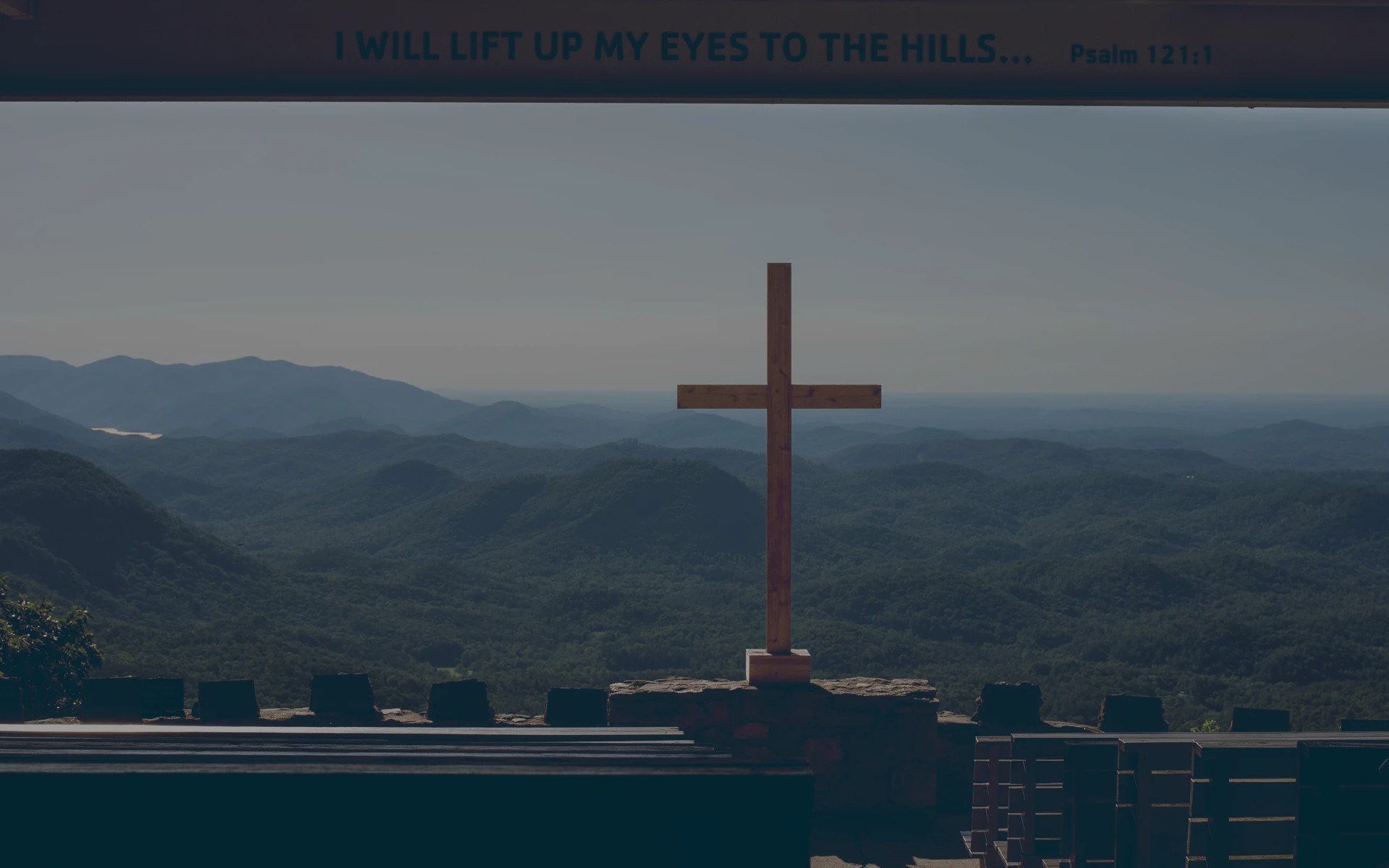 Wooden cross standing on a stone platform overlooking forested hills under a clear sky with a beam displaying the text 'I WILL LIFT UP MY EYES TO THE HILLS... Psalm 121:1'.