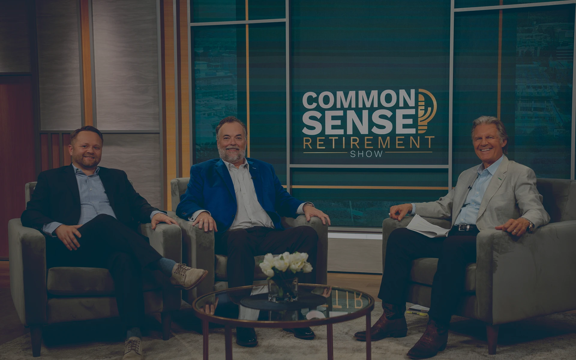 Phillip Allen, Tony Dale, and Jay Brost from Common Sense Retirement Planning sitting in armchairs on the set of the Common Sense Retirement Show with a round glass table with flowers in front of them.