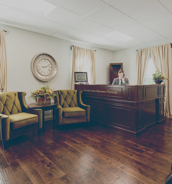Reception area with a wooden desk labeled Common Sense Retirement Planning, a receptionist on the phone, two green upholstered chairs, a round side table with a plant, and a large clock on the wall.