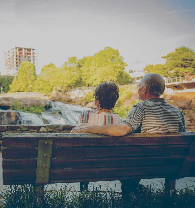 Retired couple sitting on a park bench overlooking a waterfall and greenery in an urban area.