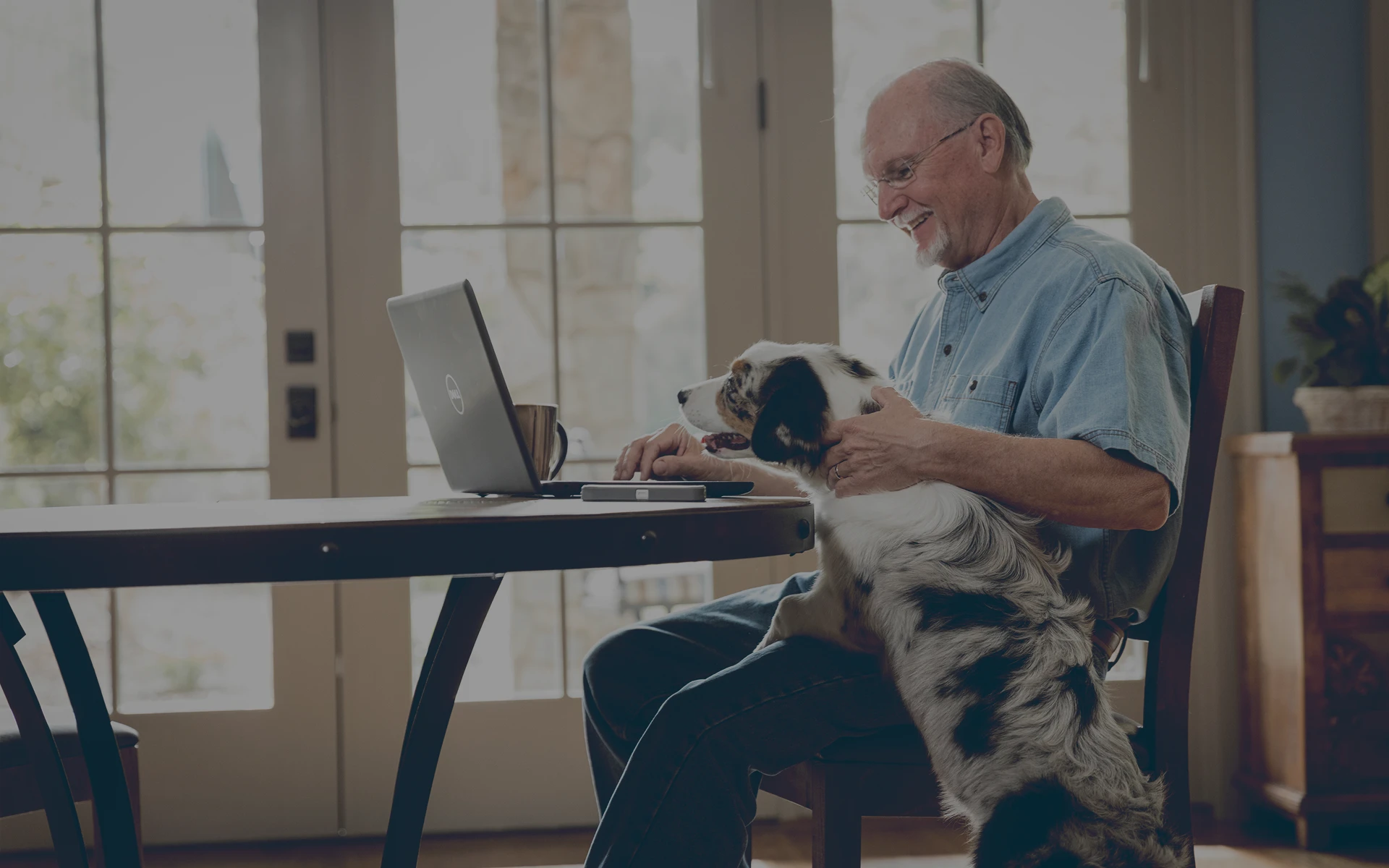 Retired man sitting at a table using a laptop while petting a black and white dog sitting beside him.