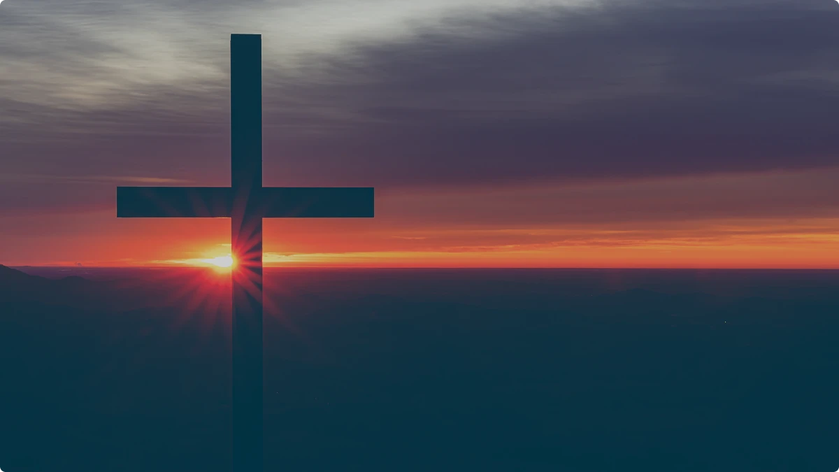 Silhouette of a cross against a vibrant sunset with sun rays and cloudy sky.