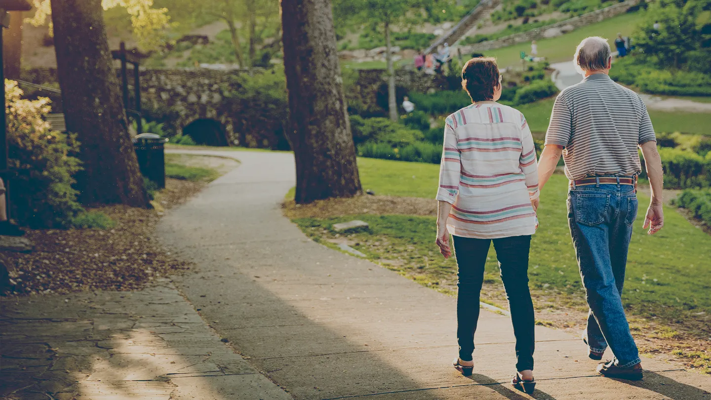 Retired couple holding hands while walking on a curved paved path in a green park during the daytime.