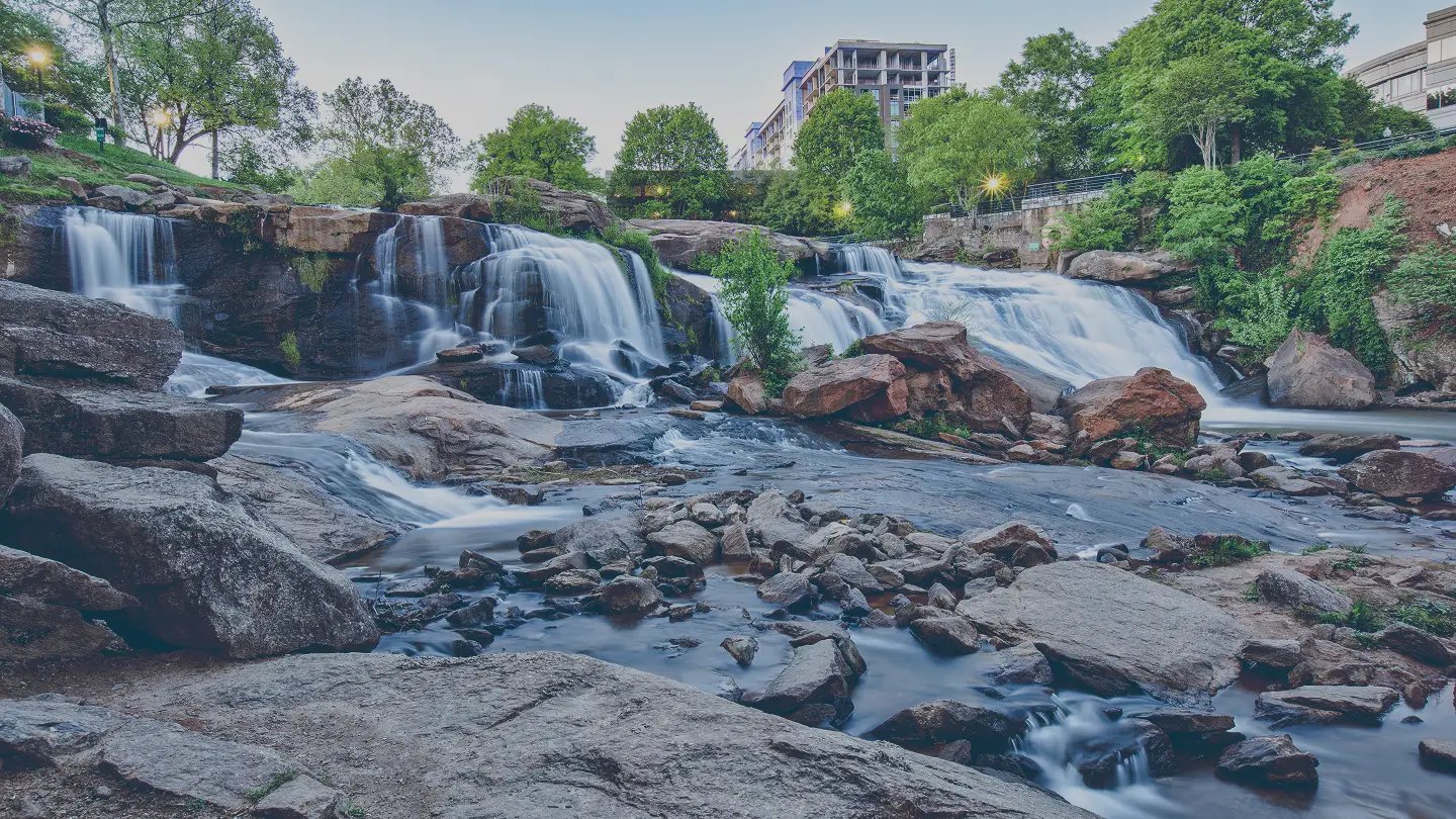 Multi-tiered waterfall flowing over rocks with greenery and buildings in the background at dusk.