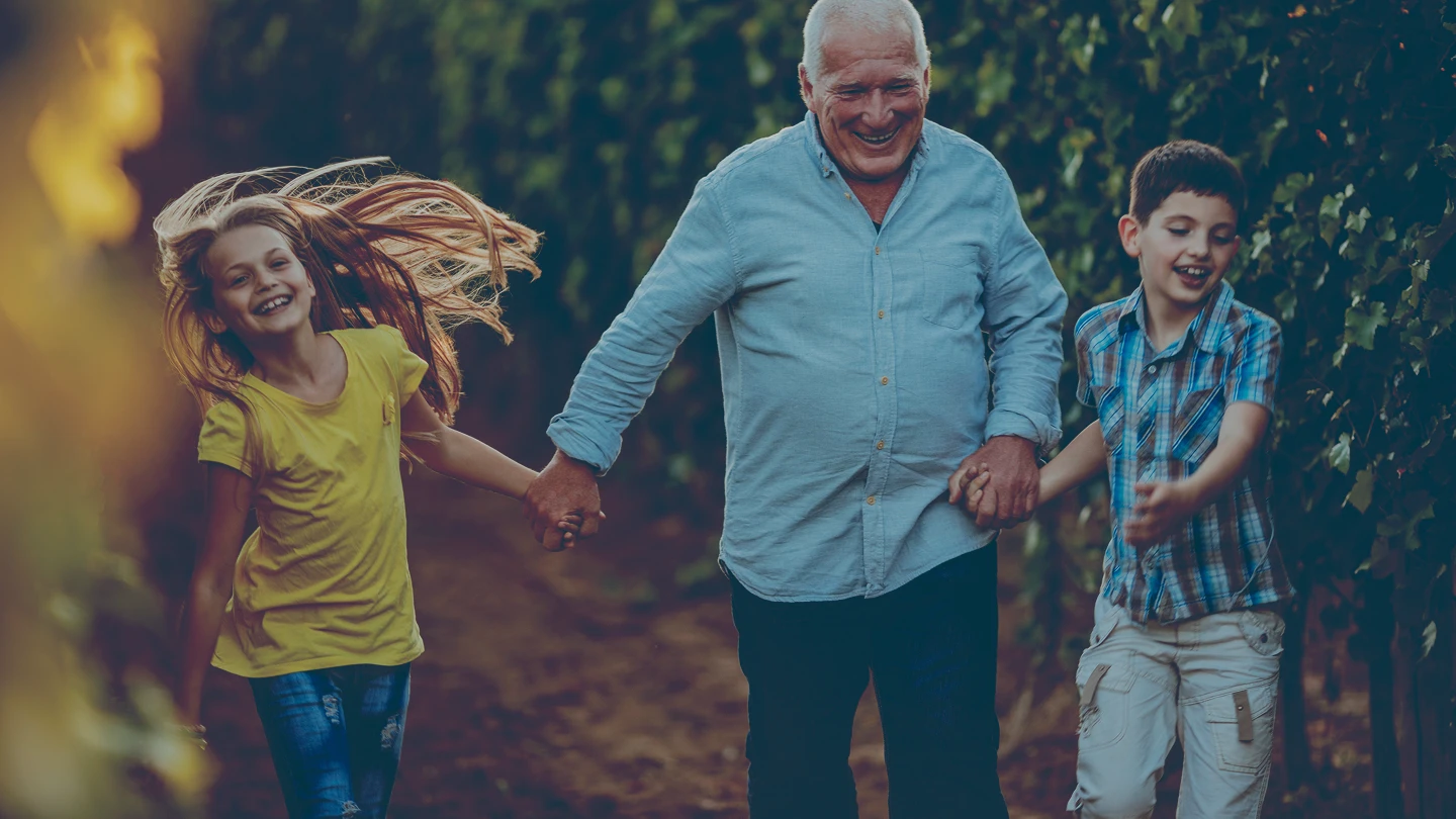 Smiling retired man holding hands and walking outdoors with two happy grandchildren, a girl in a yellow shirt and a boy in a plaid shirt.