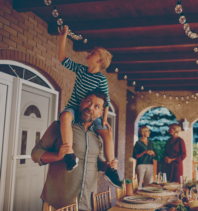 Man carrying a boy on his shoulders as the boy reaches up to touch hanging string lights in an outdoor dining area.
