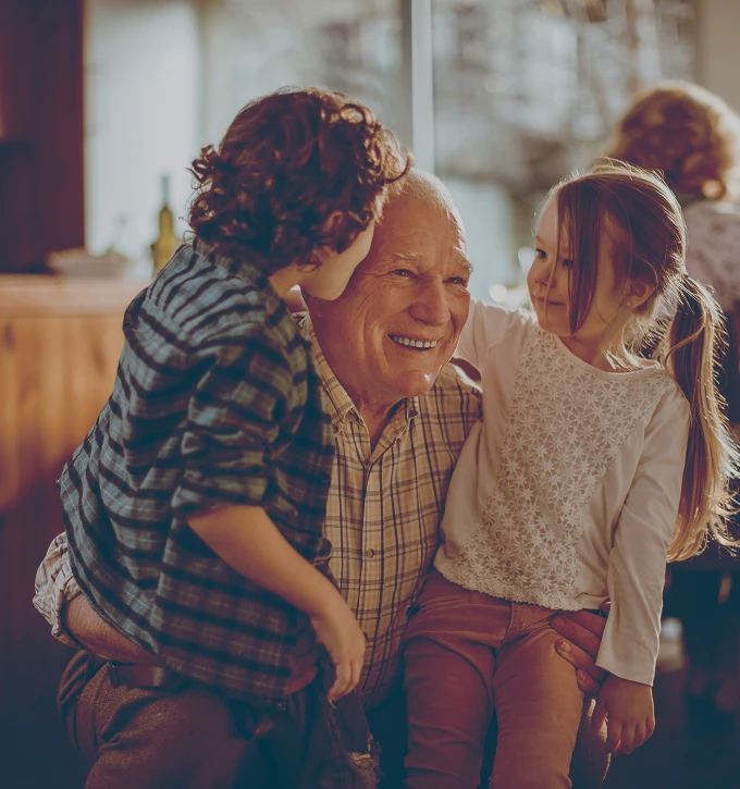 Smiling elderly man embraces two young children in a warmly lit room.