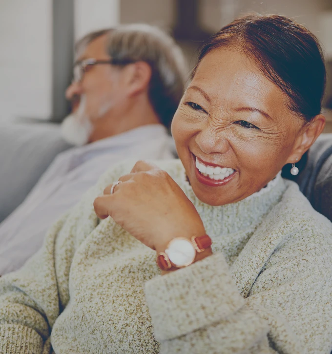 Smiling older woman in a sweater sitting on a couch with a man blurred in background.