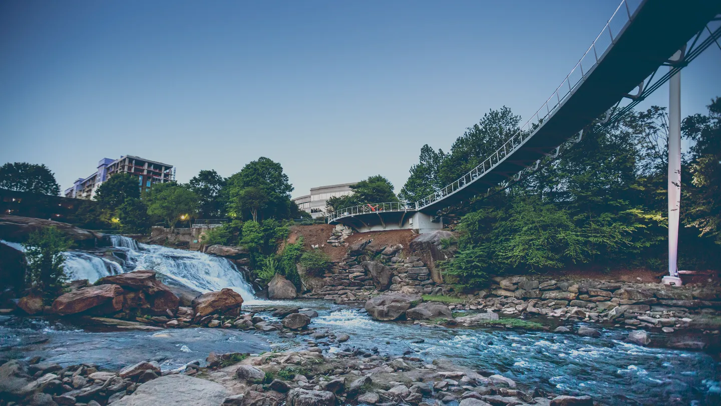 River with rocks and a waterfall under a pedestrian suspension bridge surrounded by trees and buildings at dusk.