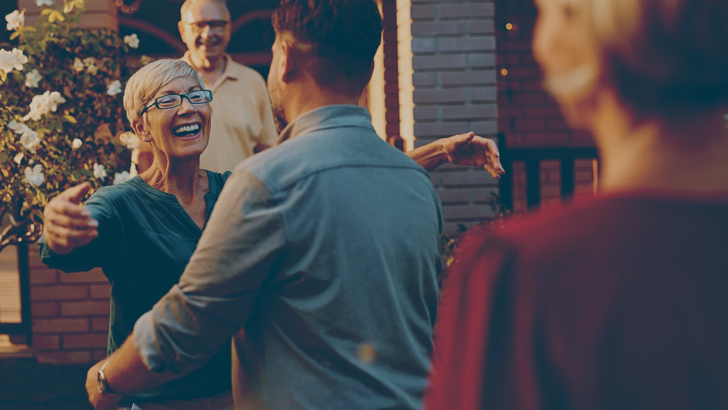 Smiling retired woman with short gray hair and glasses reaching out to hug a son, while an retired man smiles in the background near a brick house.