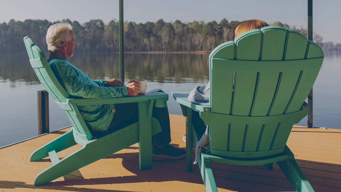Retired couple sitting in green Adirondack chairs on a wooden dock facing a calm lake surrounded by trees.