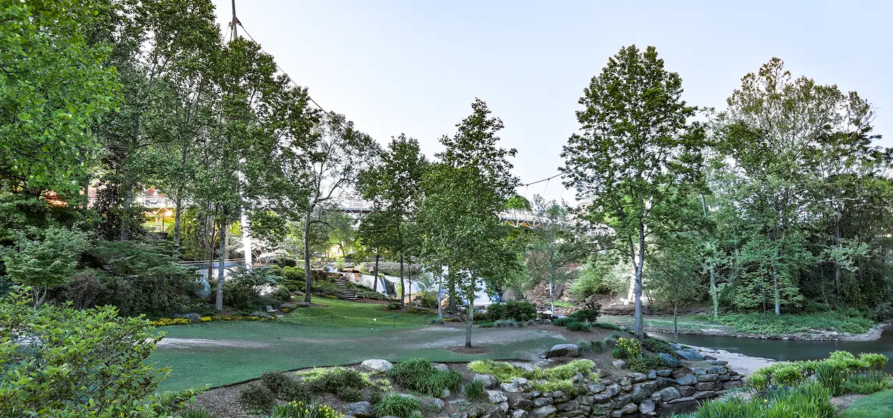 A lush park with green trees, rocks, grass, and a small river flowing beside a walking path.