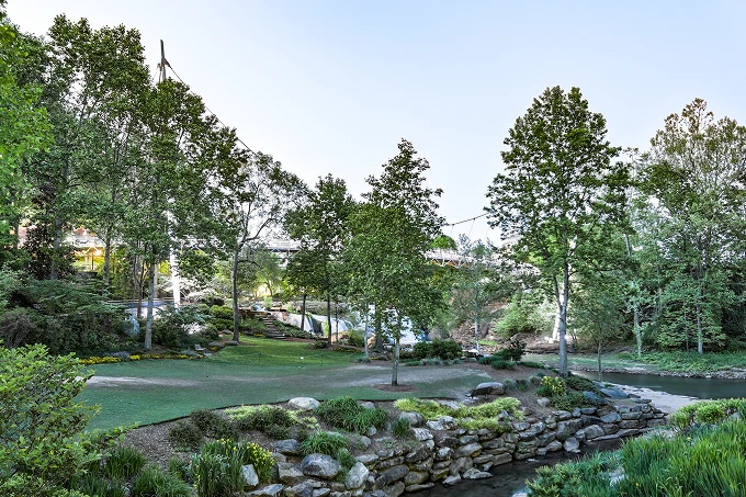 Park area with green grass, trees, rocks along a creek, and a clear sky overhead.