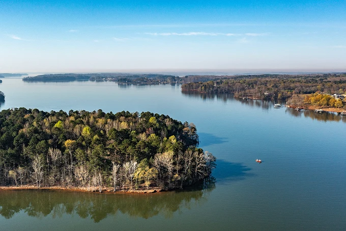 A lake with a tree-covered peninsula in the foreground and calm water extending to distant wooded shorelines.