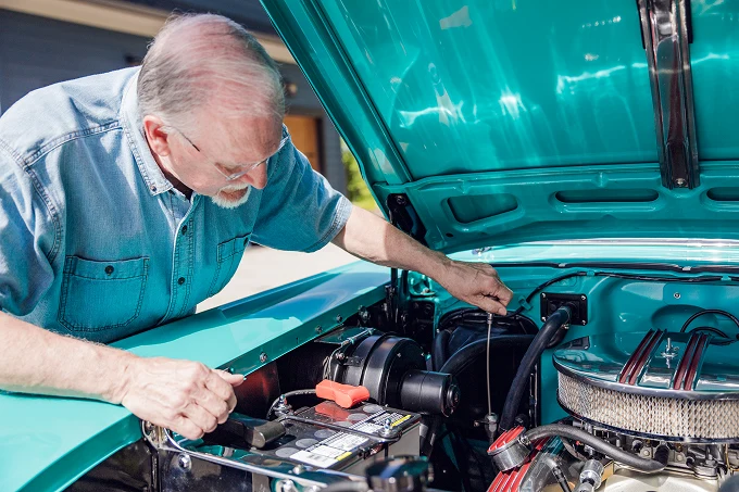 Elderly man in denim shirt checks engine oil dipstick under the open hood of a turquoise car.