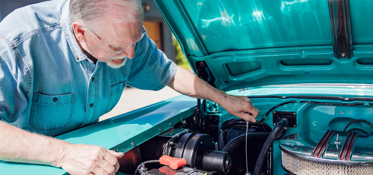 Older man in blue shirt checking engine oil dipstick under the hood of a turquoise car.